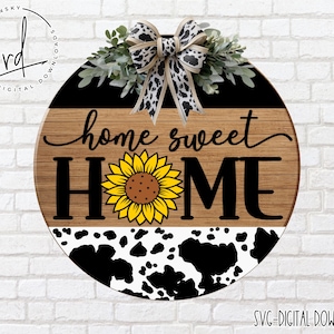 May include: A round wooden sign with a black and white cow print design, a sunflower, and the text "home sweet home".
