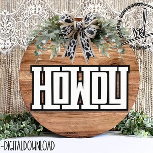 May include: A round wooden sign with the word "Howdy" in black and white letters. The sign is decorated with a brown and white leopard print bow and green leaves.