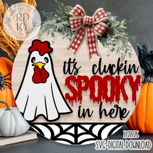 May include: A round wooden Halloween decoration featuring a ghost chicken design. The text "it's cluckin' SPOOKY in here" is in red. A red and white checkered bow and greenery are at the top. A spider web design is at the bottom.