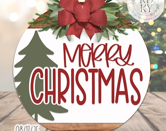 May include: A circular Christmas decoration with a white backdrop and a wooden base. The words "Merry Christmas" are in red script, with a green Christmas tree on the left. A red bow and greenery decorate the top.