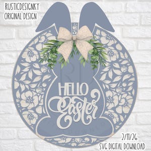 Peut inclure: Décoration de porte en bois sur le thème de Pâques, bleue et blanche, avec des oreilles de lapin et un nœud. Le design présente des découpes florales et les mots "Hello Easter". Le texte "RUSTICDESIGNKY ORIGINAL DESIGN" est en haut.