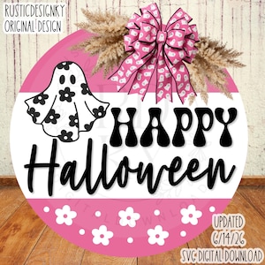 May include: A round sign featuring a pink and white background with the black text Happy Halloween. A white ghost with black flower patterns is on the left, and a pink bow with white ghost patterns sits above with dried pampas grass.