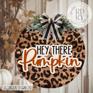 May include: Round wooden sign with a leopard print background and the text "HEY THERE Pumpkin" in white and orange. The sign is decorated with a black and white striped bow and greenery.
