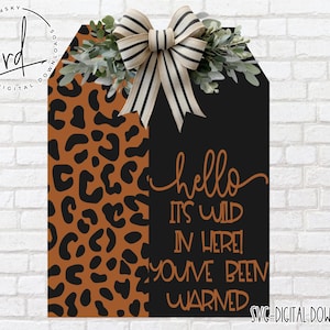 Könnte beinhalten: Ein dekoratives Holzschild mit Leopardenmuster auf der linken Seite und Schwarz auf der rechten Seite. Das Schild trägt den Text "hello it's wild in here! You've been warned" in Schreibschrift. Es ist mit einer gestreiften Schleife und Grünzeug verziert.