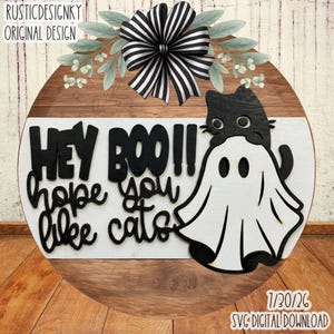 May include: A round wooden door sign featuring a black and white striped bow and green foliage at the top. The sign displays the black text HEY BOO!! hope you like cats and a graphic of a black cat wearing a white ghost sheet costume.