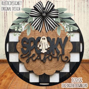 May include: A round wooden door sign featuring a black and white buffalo check pattern, a large black and white striped bow, and green leaves. The centre has a natural wood finish with the black text SPOOKY season and a white ghost icon.