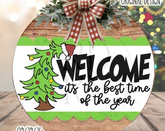 May include: A circular wooden sign with a festive Christmas design. The sign displays the word "WELCOME" in bold black letters, with the phrase "it's the best time of the year" underneath. A green Christmas tree and a red and white bow complete the design.