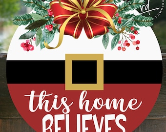 May include: A circular Christmas decoration with a Santa Claus belt design. It features a red bow with gold detailing and foliage. The text "this home BELIEVES" is in white on a red base. The words "SVG-DIGITALDOWNLOAD" are at the bottom.