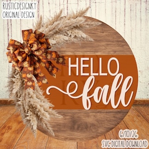 Op de afbeelding: Een rond houten bord met een horizontale oranje streep met de tekst HELLO fall in witte letters. Het bord is versierd met een grote bruine strik met een patroon van kleine oranje pompoenen en een bos beige pampasgraspluimen.