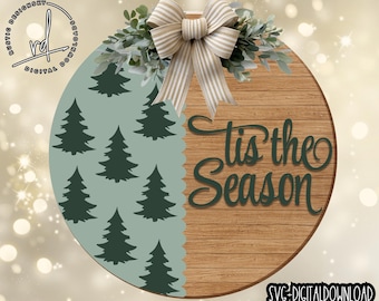 May include: A wooden round sign with a rustic wood grain finish. The sign is split in half, with the left side featuring a light blue background with a repeating pattern of dark green stylized pine trees. The right side of the sign features the text "Tis the Season" in dark green cursive lettering. The sign is decorated with a white and brown striped bow and green leaves.