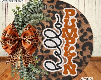 May include: A round wooden sign featuring a brown leopard print pattern and the text Hello Pumpkin in orange and white. The sign is decorated with a large orange and leopard print bow, wooden beads, and green eucalyptus sprigs on the left side.
