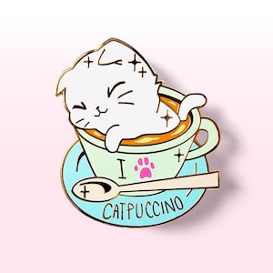 Può includere: Uno spillo in smalto dorato con un gatto bianco rannicchiato in una tazza di caffè verde e arancione con un'impronta di zampa rosa e il testo "I  CATPUCCINO".