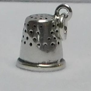 Könnte beinhalten: Ein kleiner, silberfarbener Fingerhut-Anhänger mit einer Schlaufe zum Befestigen an einer Halskette oder einem Armband. Der Fingerhut hat eine abgerundete Oberseite mit mehreren kleinen Löchern und einen leicht ausgestellten Boden. Der Anhänger ist aus glänzendem Metall.
