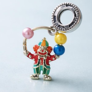 Puede incluir: Un charm de payaso colorido con un accesorio de anillo plateado. El payaso lleva una chaqueta roja, pantalones verdes y un sombrero multicolor. Sostiene una cuenta rosa y otras tres cuentas amarillas y azules.