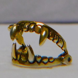 May include: A gold-colored, stylized set of teeth, likely a decorative item or jewelry. The teeth are arranged in a curved shape, with sharp, pointed canines and smaller incisors. The metal has a polished, reflective surface.