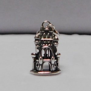 May include: A silver charm in the shape of a castle with a pointed roof, towers, and a large arched doorway.