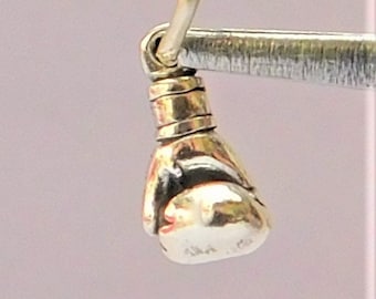 May include: A small, silver-colored boxing glove charm with a loop for hanging. The glove is detailed with a wrapped wrist and a rounded shape, held by a silver tool against a soft pink background. The charm is likely for a necklace or bracelet.