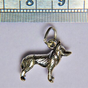 May include: Silver charm of a German Shepherd dog standing in profile. The charm has a loop at the top for attaching to a necklace or bracelet.