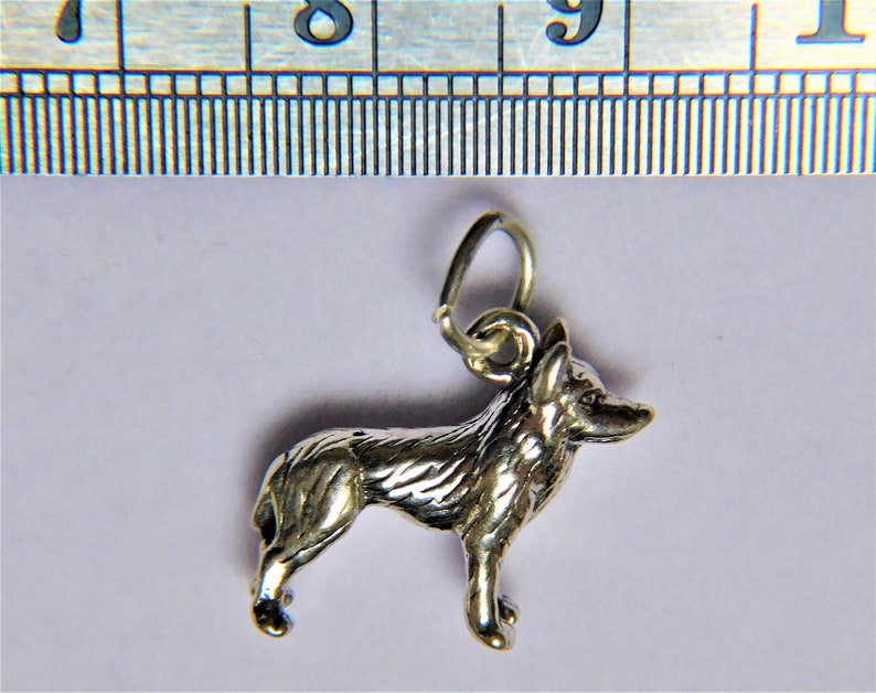 May include: Silver charm of a German Shepherd dog standing in profile. The charm has a loop at the top for attaching to a necklace or bracelet.