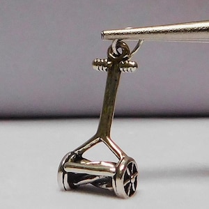 Pode incluir: Um amuleto de prata em forma de cortador de grama. O amuleto é detalhado e seria uma adição charmosa a uma pulseira ou colar.