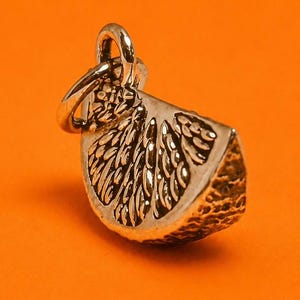 Puede incluir: Un dije de metal plateado con forma de gajo de cítrico. El dije presenta una cáscara texturizada y segmentos interiores detallados, con una pequeña argolla circular unida en la parte superior para colgarlo. La superficie tiene un acabado metálico.