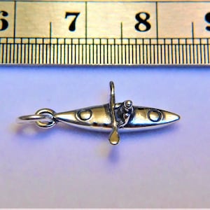May include: Silver charm of a kayak with a person paddling. The charm has a loop for attaching to a bracelet or necklace.