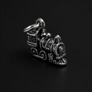 May include: A small, three-dimensional silver-toned metal charm shaped like a vintage steam locomotive train. The charm features intricate detailing on the boiler, wheels, and cab, and includes a circular jump ring attached to the top for hanging.
