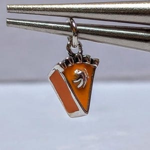 Może przedstawiać: Srebrzysty metalowy charms w kształcie kawałka ciasta dyniowego, z wypełnieniem z jasnopomarańczowej emalii i małym srebrnym kleksem bitej śmietany na wierzchu, zawieszony na małym kółeczku trzymanym przez metalową pęsetę.