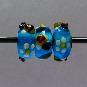 Puede incluir: Tres cuentas de vidrio con fondo azul y flores blancas con centros amarillos. Cada cuenta tiene una abeja negra y amarilla con una cara negra.