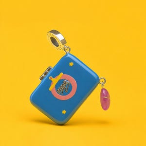 Op de afbeelding: Een blauwe emaille bedel in de vorm van een kleine koffer met een zilveren oogje. De voorkant heeft een roze cirkel, een gele clip, kleine gele sterren en de tekst MMR. Een kleine, doorschijnende roze hartvormige bedel hangt aan de zijkant.