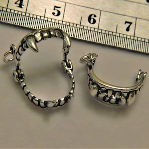 May include: Two silver-colored, detailed fang charms. One is a full circle with a hinged design, and the other is a half-circle. Both have small loops for attaching to jewelry. The charms are on a light background.