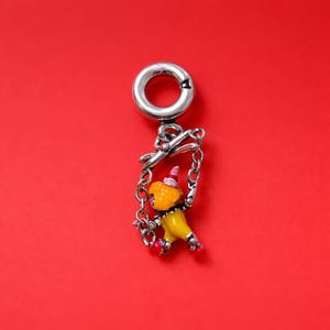 May include: A silver charm with a small, colorful clown figure swinging from a chain. The clown has a yellow outfit, a pink hat, and red shoes. The charm has a clasp at the top and is set against a red background.