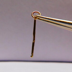 Könnte beinhalten: Ein goldfarbener Häkelhaken, der von einer silberfarbenen Zange gehalten wird. Der Haken hat oben eine kleine Schlaufe und ein gebogenes Ende. Das Werkzeug wird zum Häkeln verwendet.