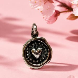 May include: A silver-toned metal charm shaped like a wax seal, featuring a raised heart icon in the center surrounded by a ring of small dots. The charm has an irregular, organic edge and includes a small circular loop at the top for attachment.
