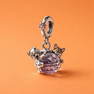 May include: A silver charm with two cat figures hugging a faceted purple sphere. The charm has a loop at the top with a pink gemstone. The cats have gold and pink accents.