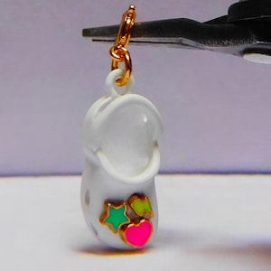 Pode incluir: Encanto branco em forma de tamanco com um laço dourado no topo. O tamanco tem três decorações coloridas: uma estrela verde, uma estrela verde limão e um coração rosa.