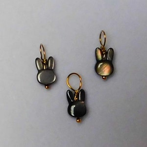 May include: Three black and iridescent shell bunny charms with gold wire loops. The charms are hanging from a white background.