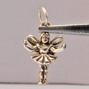 May include: A silver charm in the shape of a fairy with wings, a dress, and a smiling face.
