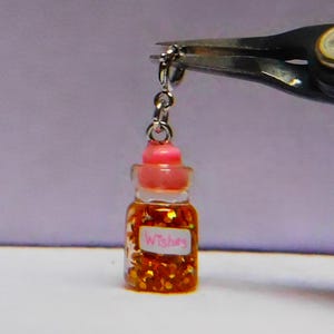 Pode incluir: Uma pequena garrafa de vidro cheia de glitter dourado e uma tampa rosa. A garrafa tem um rótulo branco que diz "Wishes" em letras cor-de-rosa. A garrafa está presa a uma corrente prateada.