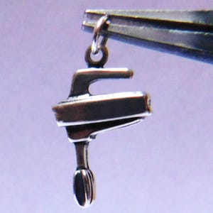 May include: A small, silver-colored charm in the shape of a hand mixer. The charm is suspended from a silver ring and held by a pair of pliers. The mixer has a detailed design, including the handle and the beater.