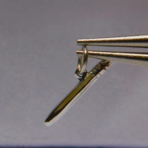 Op de afbeelding: Close-up van sieradenmaakgereedschap dat een kleine, zilverkleurige hanger vasthoudt. De hanger heeft een slanke, langwerpige vorm met een afgeronde punt. De gereedschappen zijn zilverkleurig en de achtergrond is zachtgrijs.