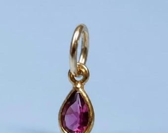 May include: A small gold pendant featuring a teardrop-shaped, faceted magenta gemstone set in a polished gold bezel. The pendant includes a circular gold jump ring at the top, designed for attachment to a necklace chain or bracelet.