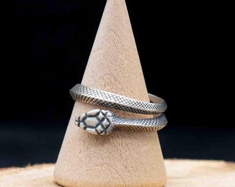 May include: A silver-colored snake ring with a textured pattern wraps around a wooden cone. The snake's head features a detailed design. The ring is displayed on a small wooden slice against a dark background.