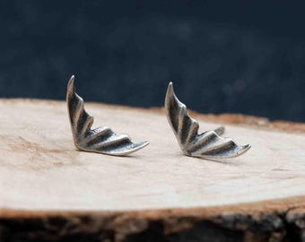 May include: A pair of silver-colored stud earrings, each shaped like a stylized wing. The earrings are displayed on a rough-hewn wooden surface against a dark background. The wing design features detailed lines, adding texture to the metal.