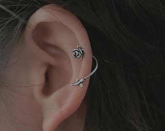 May include: Close-up of an ear featuring two pieces of silver jewellery. A small rose stud sits in the upper ear, and a silver ear cuff with a floral design curves along the ear. Dark hair frames the ear.