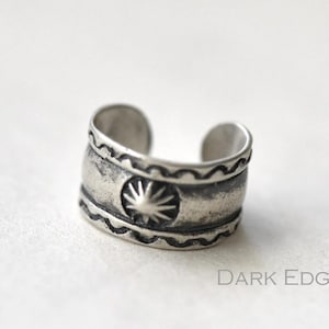 May include: A silver-toned ear cuff with an open design. The cuff features a sunburst design in the center and decorative patterns along the edges. The text "Dark Edge" is visible.