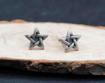 May include: A pair of silver-toned star-shaped stud earrings. Each earring features a five-pointed star design with interwoven lines, creating a textured appearance. The earrings are displayed on a light-colored wooden surface.