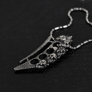 May include: A silver pendant necklace with a claw-like design. The pendant has a textured surface with skull details and circular cutouts. The necklace chain is made of small, round links.