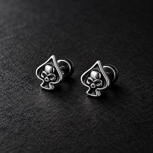May include: A pair of silver-toned stud earrings shaped like the spade suit from a deck of cards. Each earring features a raised skull design centred within the spade outline, with a polished metallic finish and a standard post backing.