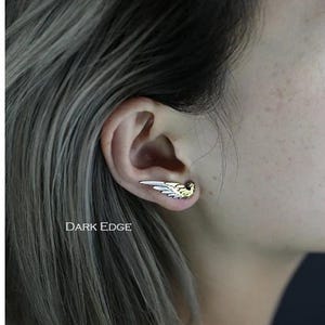 May include: A silver and gold wing earring with a delicate design. The earring is worn in the upper lobe of the ear.